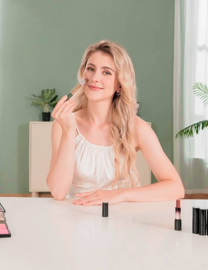 Makeup artist applying blush to woman sitting at a table with cosmetics products.