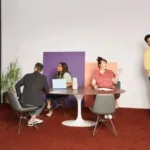 Group of professionals collaborating around a table with the Whiteboard Owl video conferencing device in a modern office setting, featuring vibrant wall colours and a relaxed atmosphere.